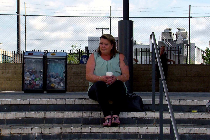 Fran sits on the steps of a train station holding a coffee.