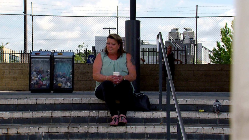 Fran sits on the steps of a train station holding a coffee.