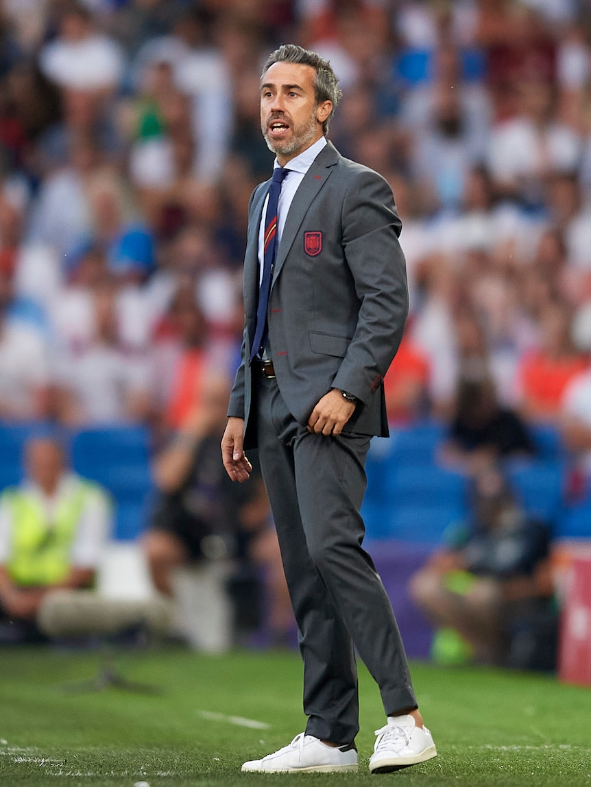 A man wearing a grey suit walks on a soccer pitch with a big crowd in the background