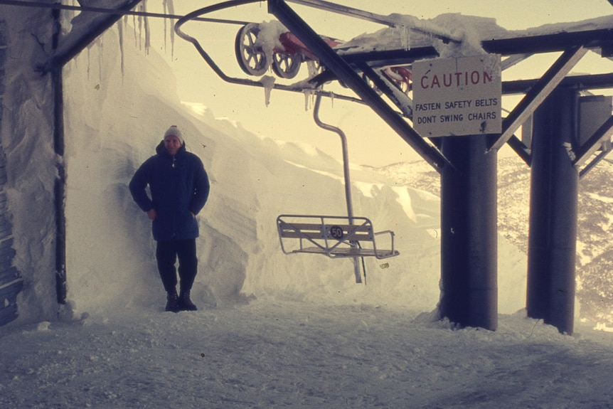 Person in snow gear stands by ski chair lift in many metres of snow