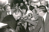 Black and white photo of Ewart holding a microphone in media pack filming Fraser getting into a car.