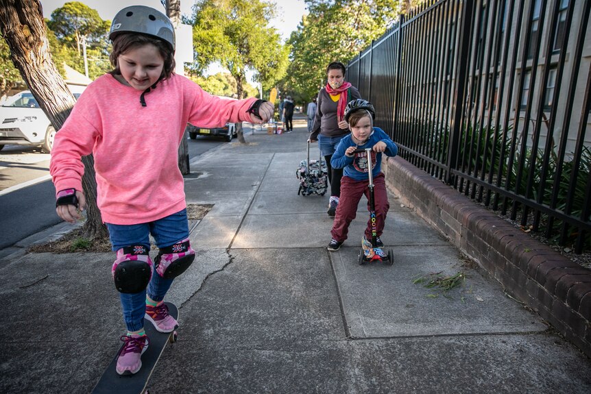Elena wheels a shopping cart behind her as her children skateboard and scoot home.