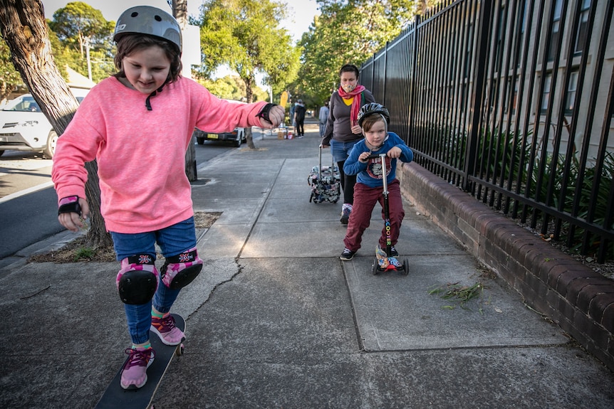 Elena wheels a shopping cart behind her as her children skateboard and scoot home.