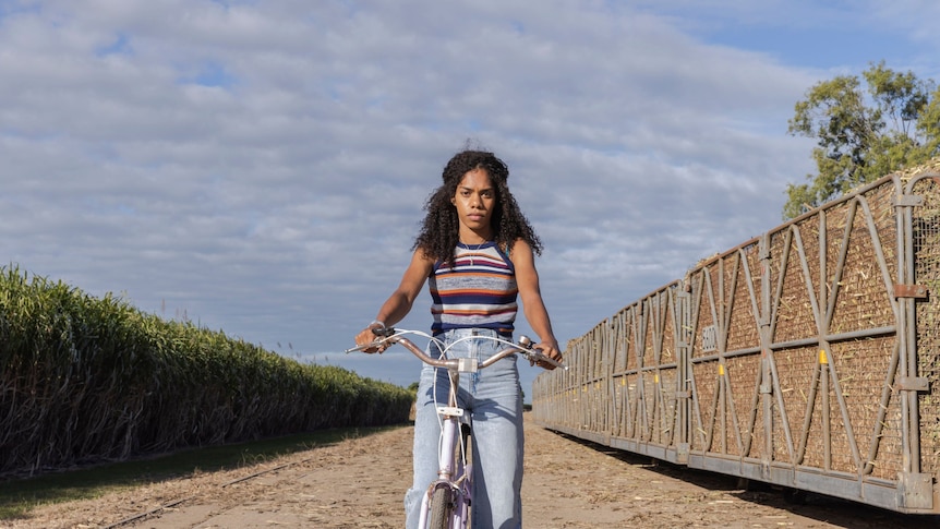 A young South Sea Islander woman riding a bike between a cane train and cane field. 