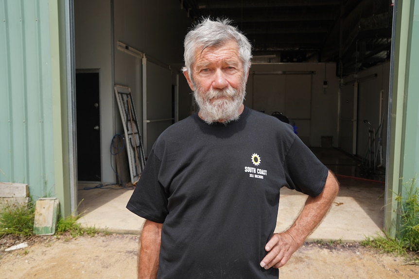 A man with a grey beard looks at a camera with a hand on his hip.