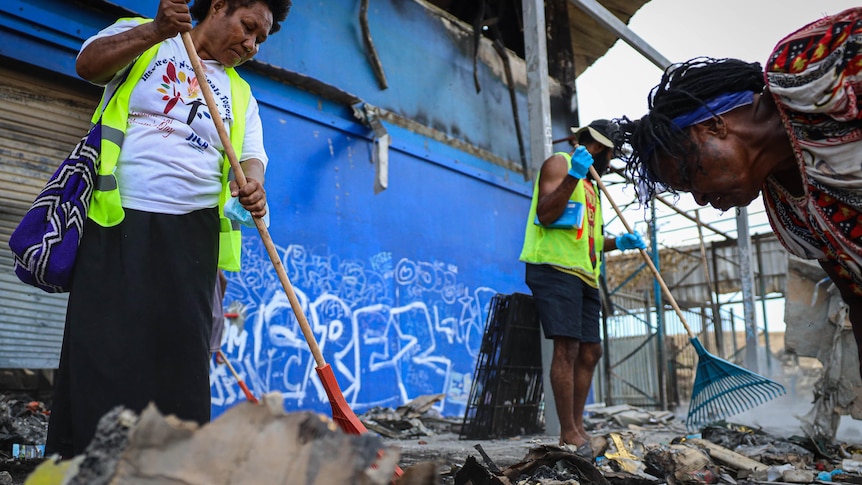 Three people hold brooms as they sweep debris off the ground in front of a store.