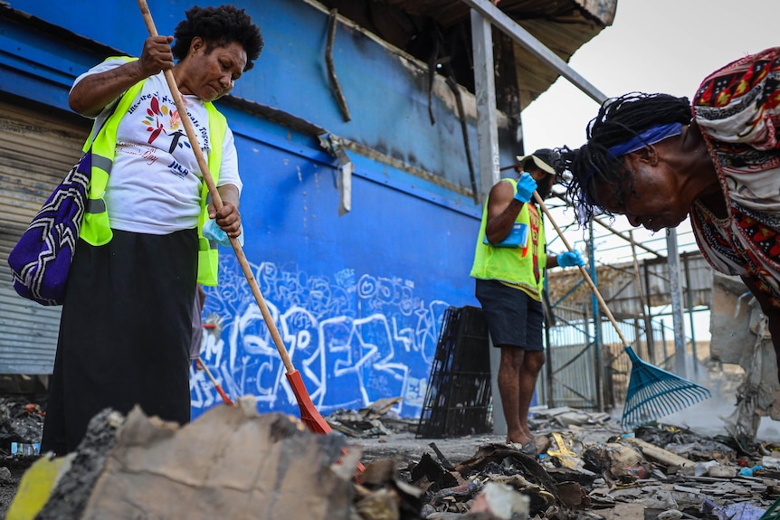 Three people hold brooms as they sweep debris off the ground in front of a store.