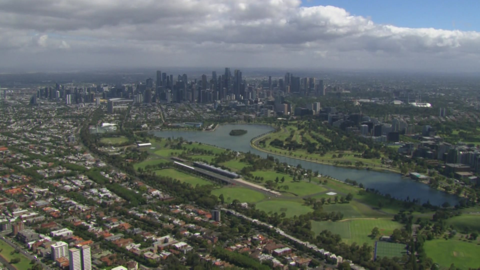 An aerial shot of a lake surrounded by green fields and houses with the city skyscrapers in the distance.