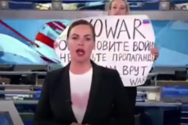 A person holds a sign reading "no war" during a live studio news broadcast.