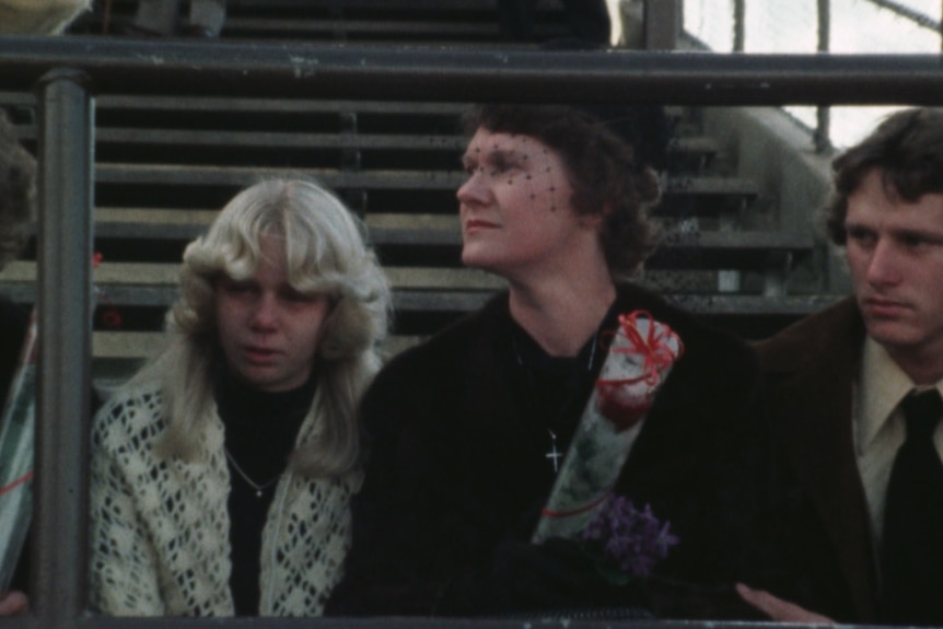 Woman dressed in black clothes and veil sits in sportsground next to a tearful teenage girl.