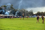 A player kicks a football over the man on the mark as other players and spectators watch on at a country footy ground 