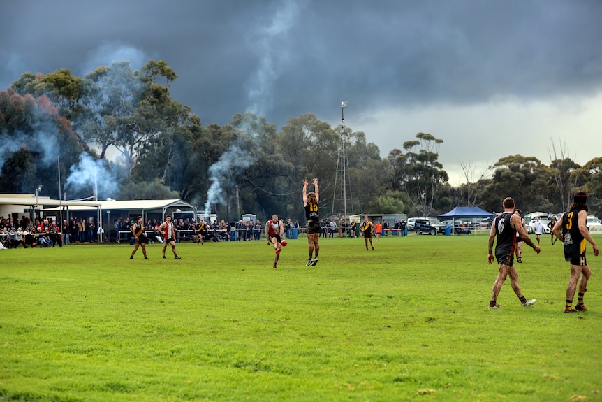 A player kicks a football over the man on the mark as other players and spectators watch on at a country footy ground 