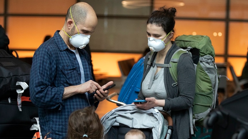 A family checking their mobile phones at an airport, both parents wear face masks.
