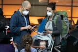 A family checking their mobile phones at an airport, both parents wear face masks.