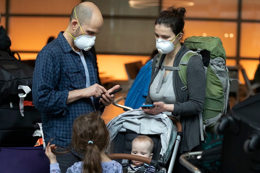 A family checking their mobile phones at an airport, both parents wear face masks.