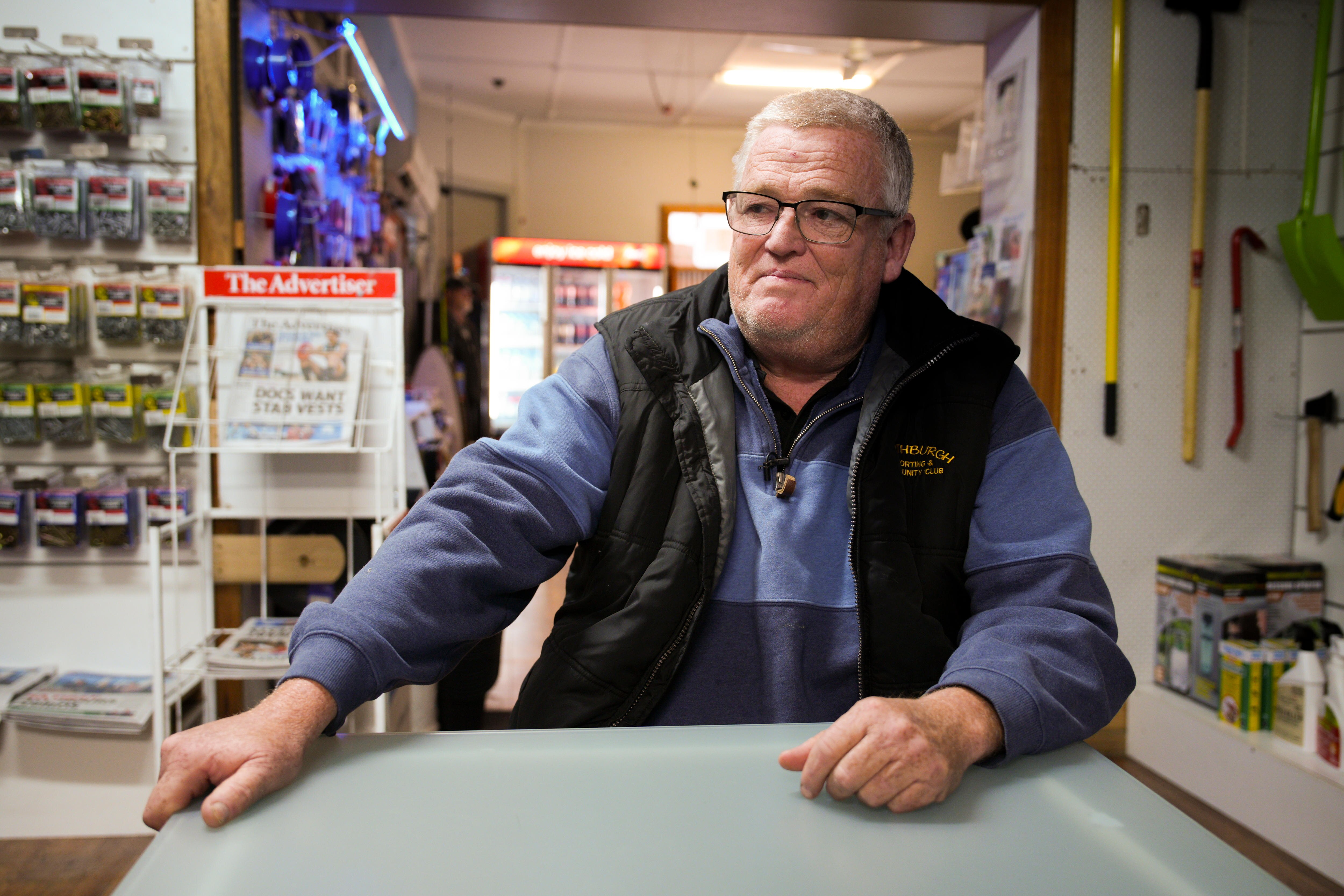 Un hombre usa un jersey azul, se ve solemnemente fuera de la cámara, sus manos se apoyan en una tienda. Detrás de él hay bienes para la venta