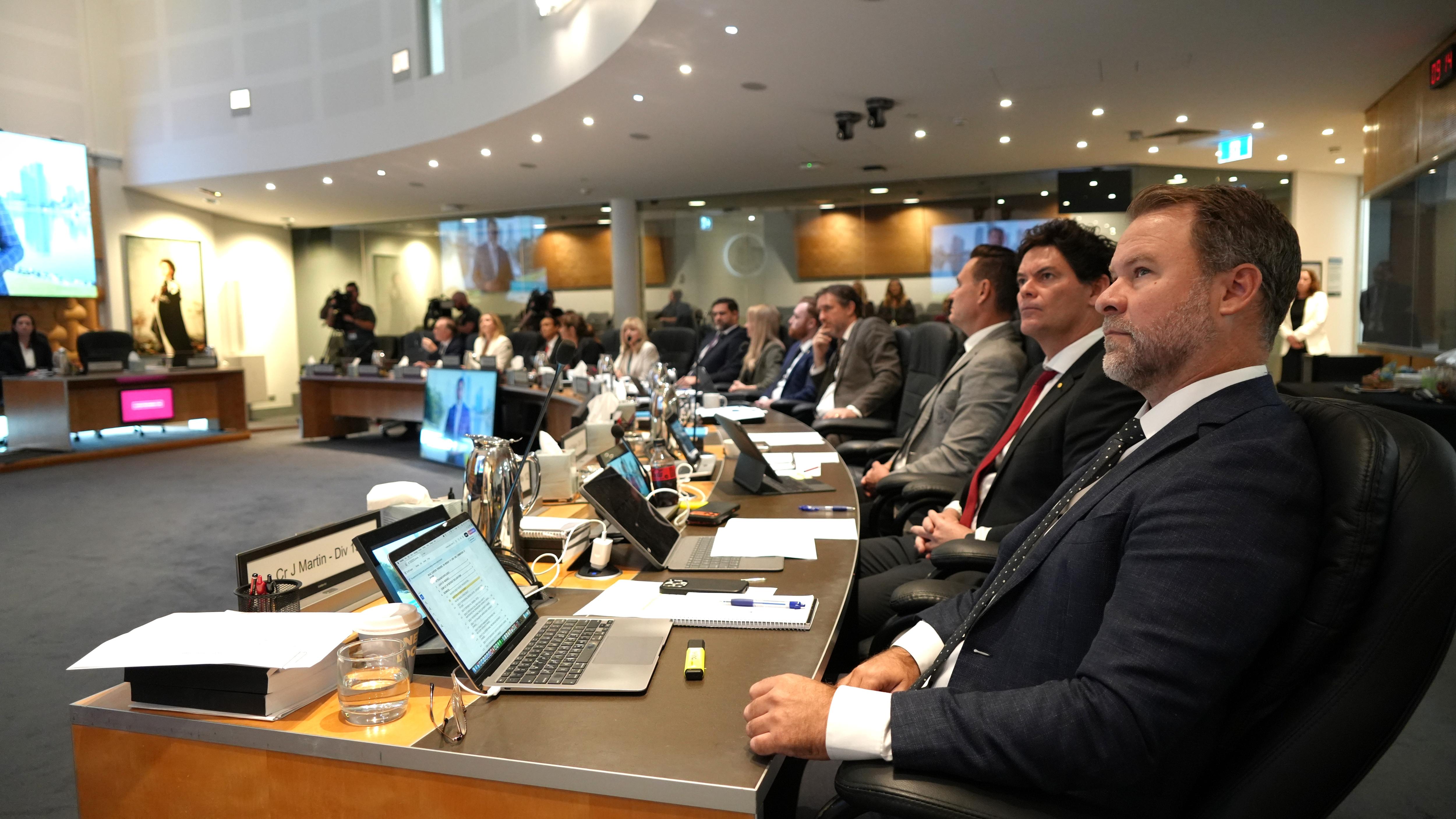 Formally dressed men and women sit at a long, curved table in a council chamber.