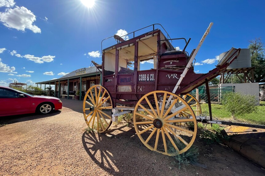 An old red wooden carriage with white wheels sits in the sun in front of an old outback pub.