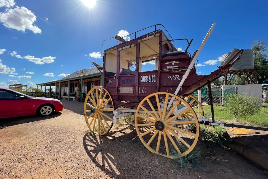 An old red wooden carriage with white wheels sits in the sun in front of an old outback pub.