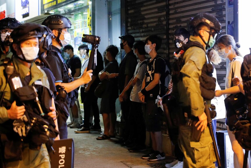 Riot police wearing masks line protesters up against a wall.