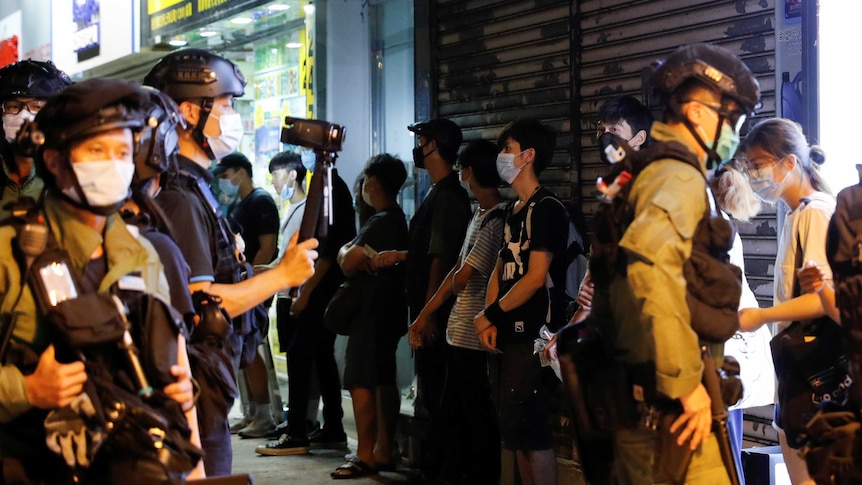 Riot police wearing masks line protesters up against a wall.