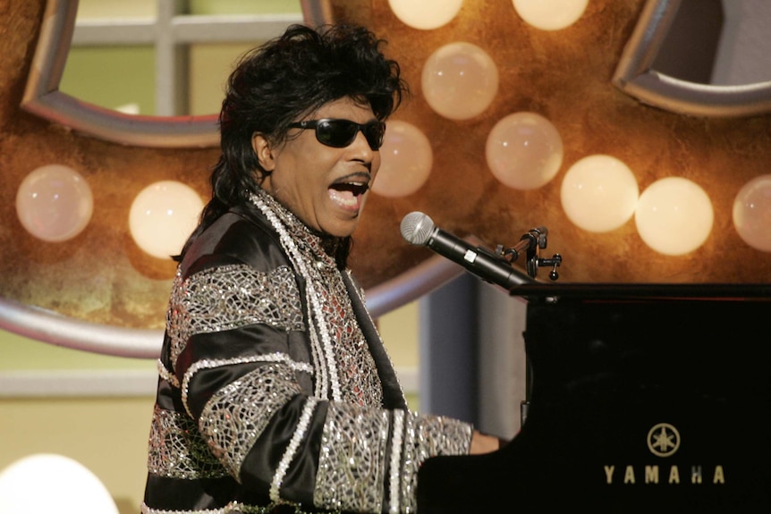 A man wearing a sparkly jacket plays at a piano while singing into a microphone.