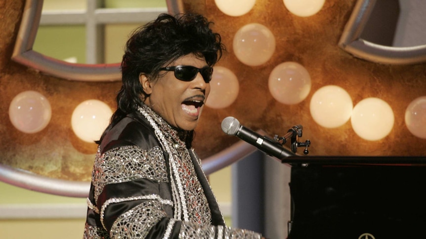 A man wearing a sparkly jacket plays at a piano while singing into a microphone.