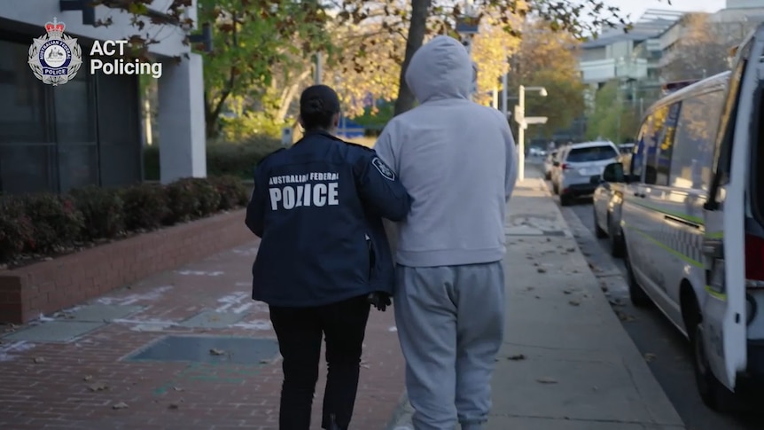 A police officer walks beside a taller individual in a grey tracksuit along a street next to a police van.