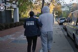 A police officer walks beside a taller individual in a grey tracksuit along a street next to a police van.