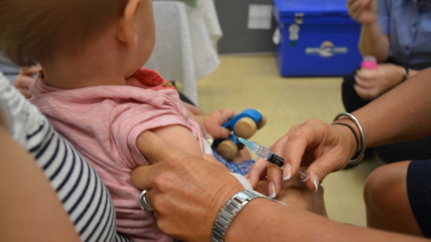 A nurse prepares to give a needle to a baby being held by a woman in a dimly-lit hospital room.