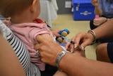 A nurse prepares to give a needle to a baby being held by a woman in a dimly-lit hospital room.