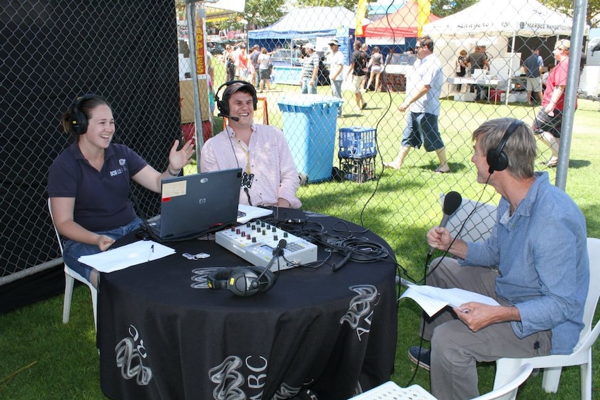 Broadcasters with headphones on sitting around broadcasting equipment on a table at an outdoor market.