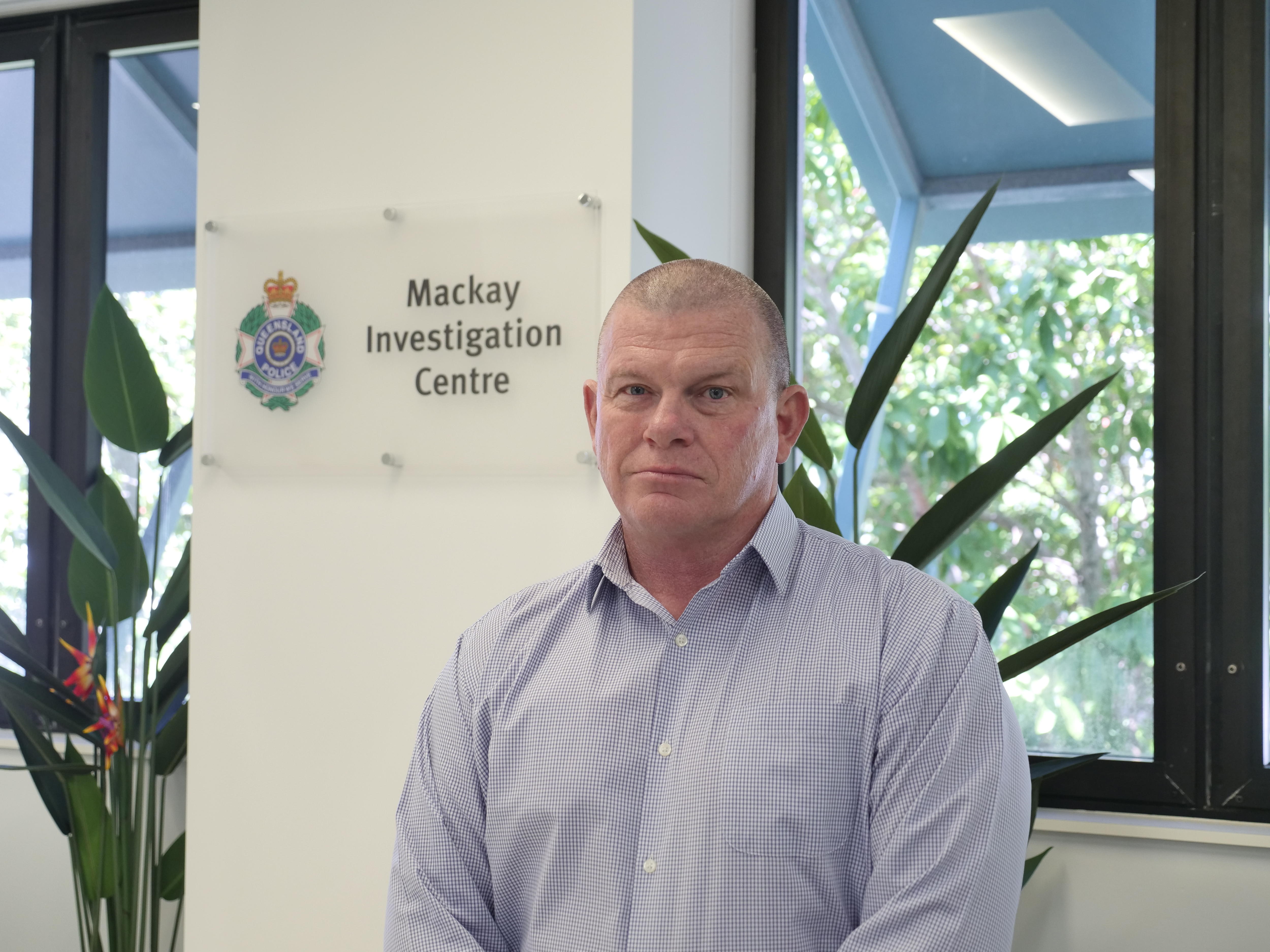 Um homem de meia-idade, com a cabeça raspada e uma camisa de botão, está em frente a uma placa da polícia.