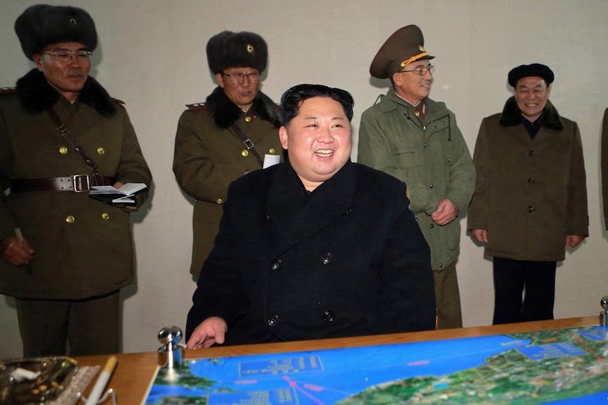 North Korean leader Kim Jong-un sits at desk with map on it and generals standing behind him