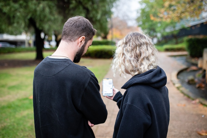 Couple face away from the camera and look at a phone in the middle of them. 