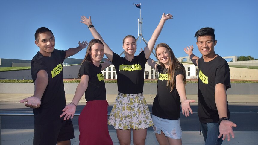 Five young people in Heywire shirts stand in front of Parliament House with their arms in the air.