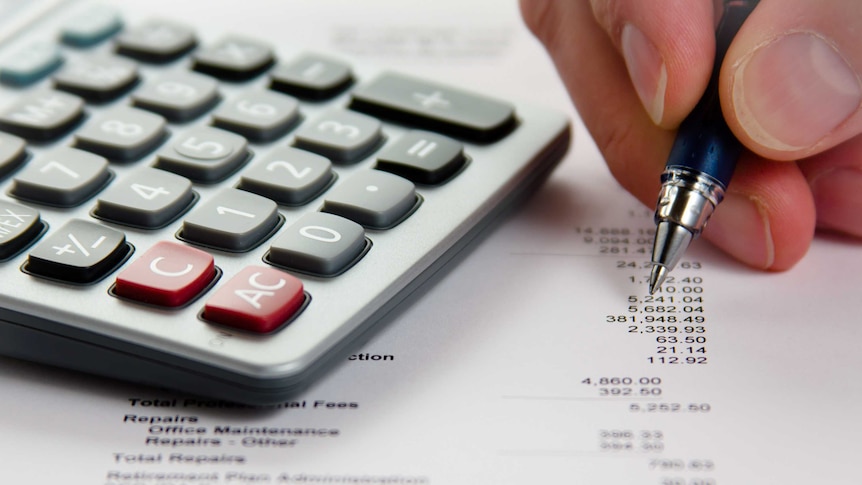 A calculator next to a hand holding a pen on a paper with numbers