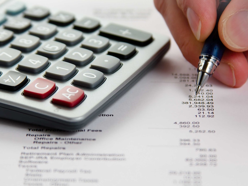 A calculator next to a hand holding a pen on a paper with numbers