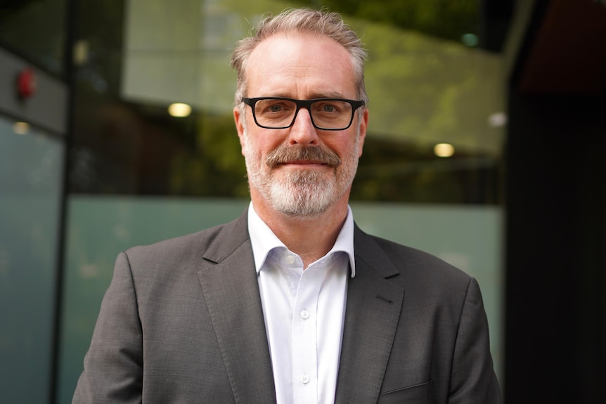 A man with a beard wearing black glasses, a grey suit jacket and button down shirt smiles at camera. 