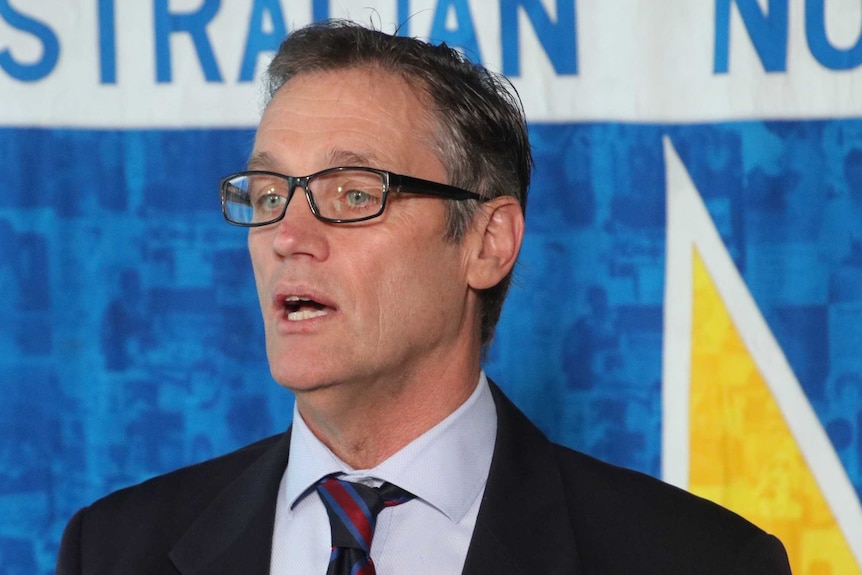 Mark Olsen wearing a suit at a press conference in front of a nurses union sign.