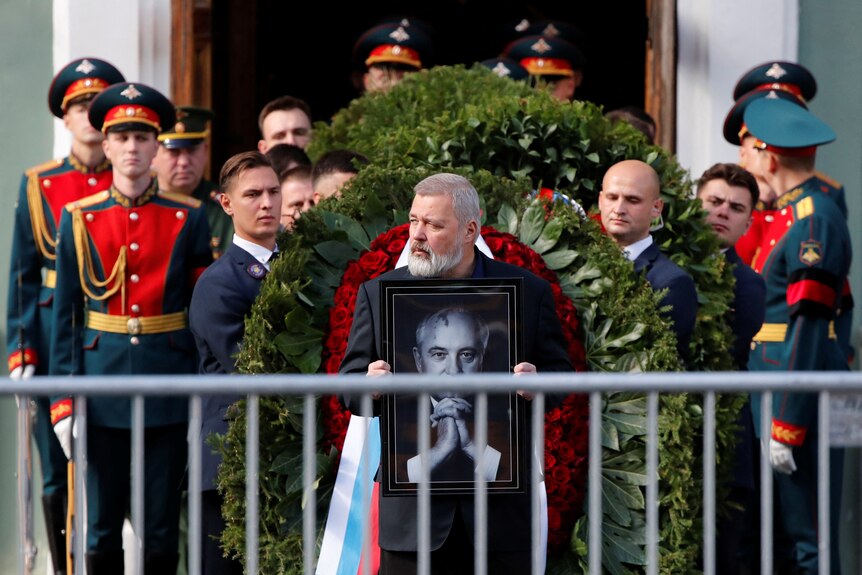 Bearded man holds portrait in front of funeral procession.