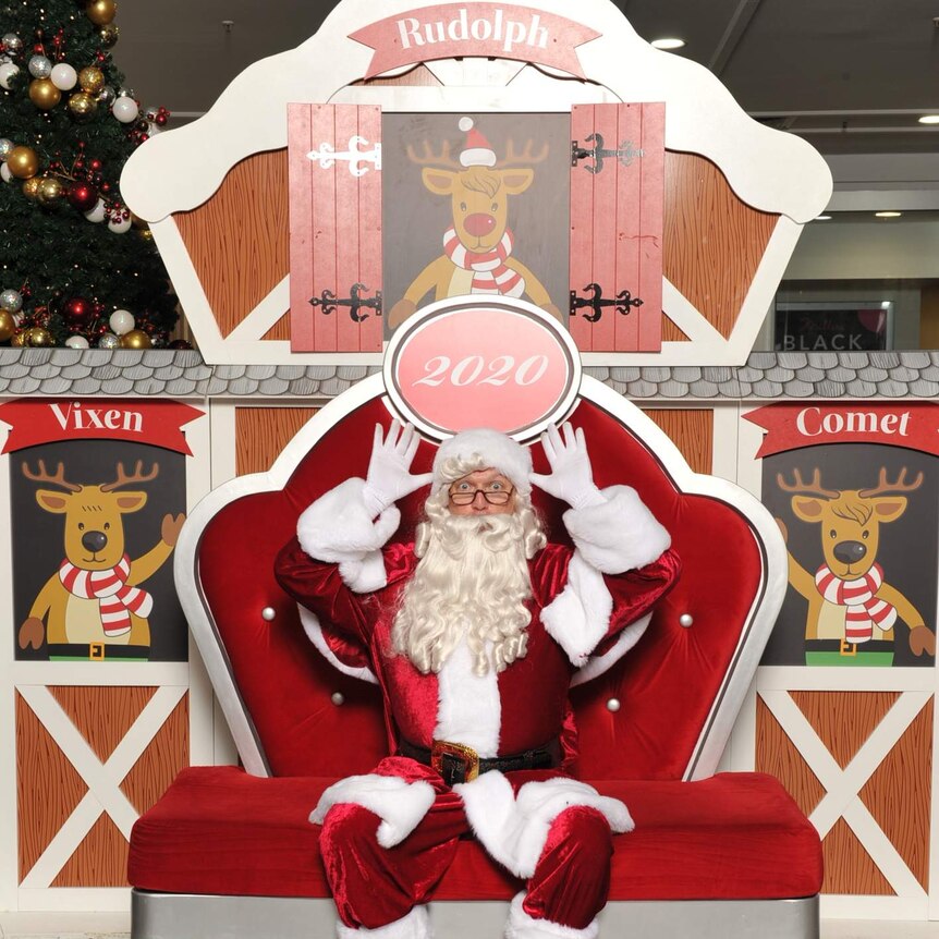 Glen Phillips wears a Santa suit and sits on a plush red seat. He has his hands placed near his ears, like antlers.