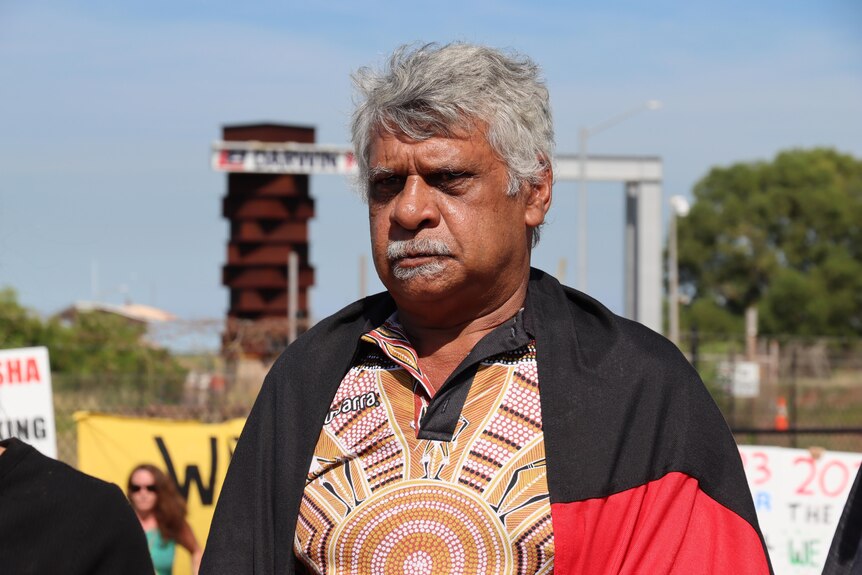 An Indigenous man looking serious and standing outside on a sunny day, with port infrastructure in the background.