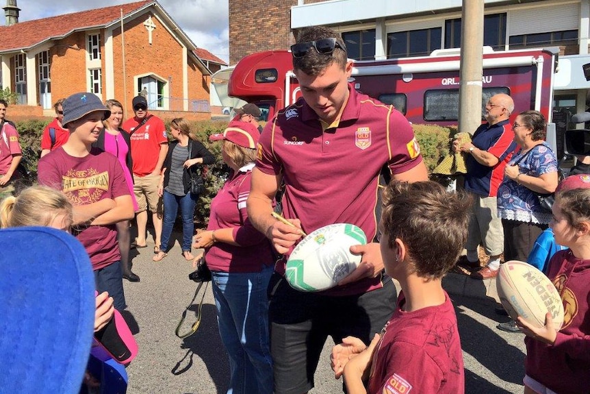 A man in a maroon shirt signing a football, surrounded by children, with buildings and a bus in the background.
