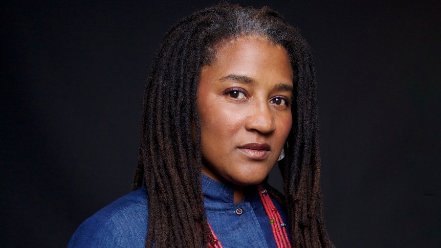 A headshot of Lynn Nottage, a black woman with dreadlocks, in front of a dark backdrop looking seriously at the camera.