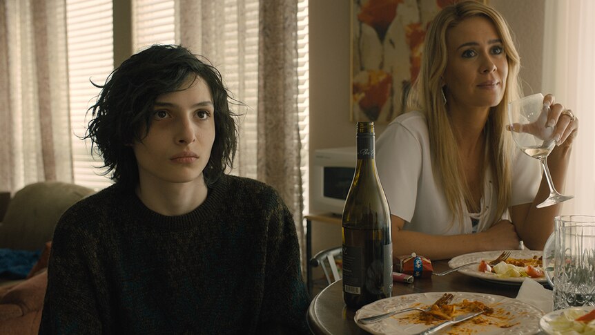 A raven-haired pale young boy sits at small dining room table with blonde-haired woman drinking wine.