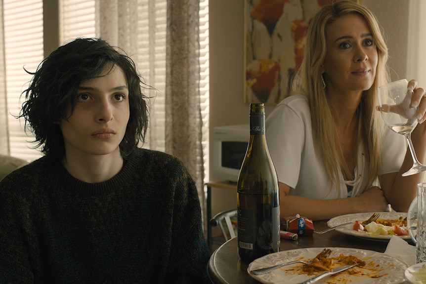 A raven-haired pale young boy sits at small dining room table with blonde-haired woman drinking wine.