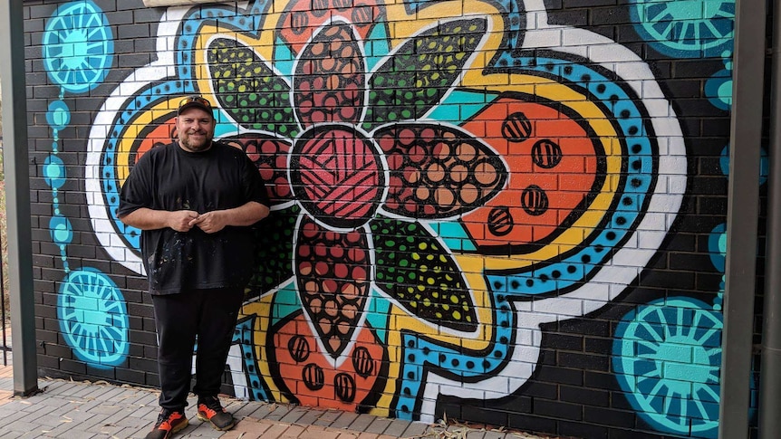 A man wearing black pants and a black t-shirt spattered with paint stands in front of a flower mural on a brick wall.
