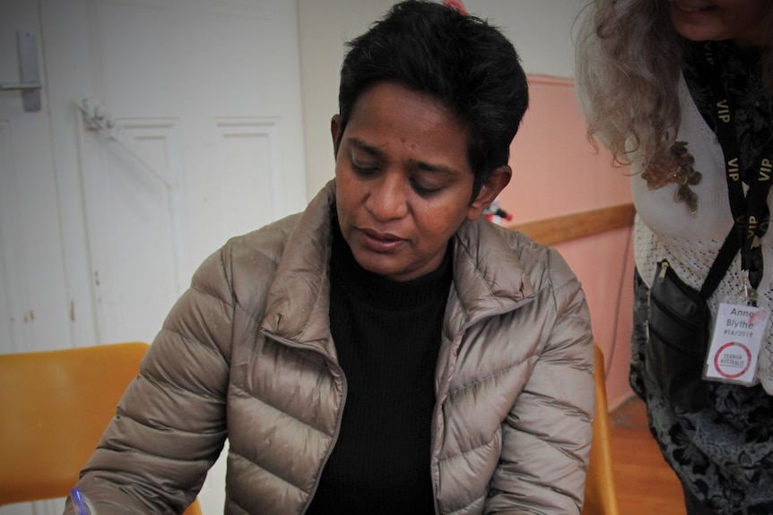 A middle aged Indian female in grey puffer jacket sits and signs a book