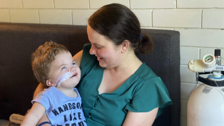 A woman holds a little boy who is smiling. He has medical tape on his cheek